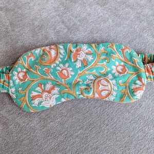 May include: A teal sleep mask with an elastic band, featuring a floral pattern in orange, white, and gold. The mask is designed to block out light for better sleep. The floral design is repeated across the mask.
