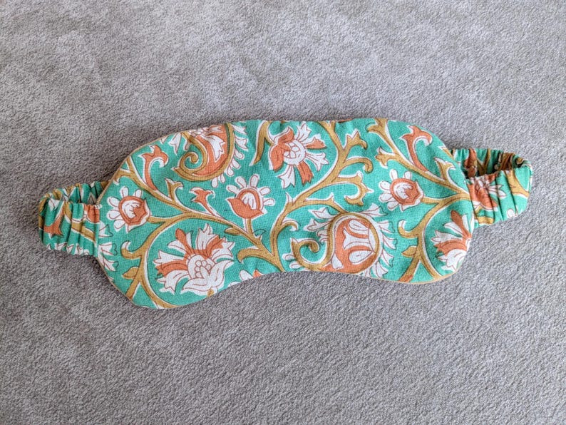May include: A teal sleep mask with an elastic band, featuring a floral pattern in orange, white, and gold. The mask is designed to block out light for better sleep. The floral design is repeated across the mask.