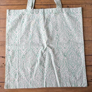 May include: A white tote bag with a repeating light teal damask pattern. The bag has two matching teal handles and is made of a soft fabric. The bag is sitting on a wooden surface.