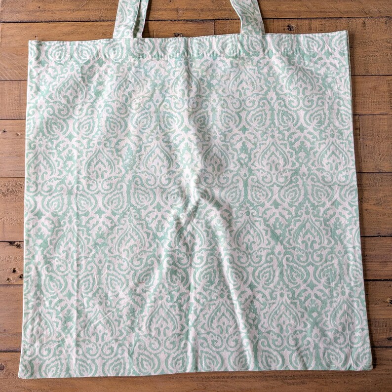 May include: A white tote bag with a repeating light teal damask pattern. The bag has two matching teal handles and is made of a soft fabric. The bag is sitting on a wooden surface.