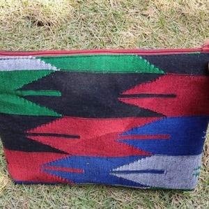 May include: A rectangular, multi-coloured zipped pouch with a geometric pattern. The design features alternating bands of green, black, red, blue, and grey. The zip and trim are red. The pouch is resting on grass.