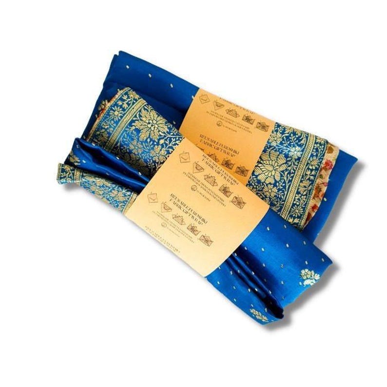 May include: A blue silk scarf with gold and white floral patterns is wrapped around a smaller blue silk scarf with white polka dots. The scarves are packaged with a brown paper label that includes instructions for different ways to wear the scarf.