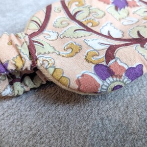 May include: Close-up of a floral patterned headband. The fabric features a peach background with a repeating design of purple, yellow, and white flowers and leaves, outlined in burgundy. The elasticated section is gathered and visible.