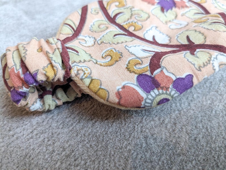 May include: Close-up of a floral patterned headband. The fabric features a peach background with a repeating design of purple, yellow, and white flowers and leaves, outlined in burgundy. The elasticated section is gathered and visible.