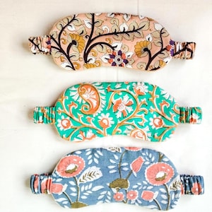 May include: Three patterned sleep masks. The top mask has a peach background with a floral design in brown, yellow, and purple. The middle mask is green with orange and white floral patterns. The bottom mask is blue with pink flowers.
