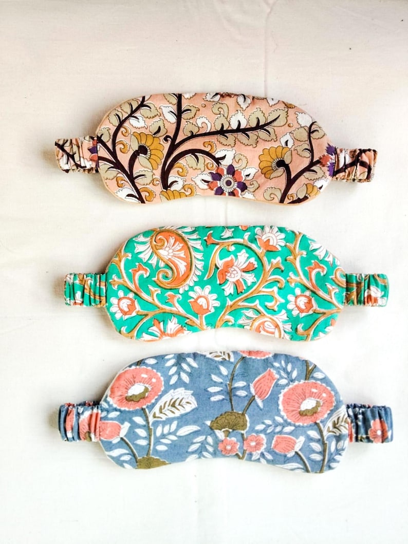 May include: Three patterned sleep masks. The top mask has a peach background with a floral design in brown, yellow, and purple. The middle mask is green with orange and white floral patterns. The bottom mask is blue with pink flowers.