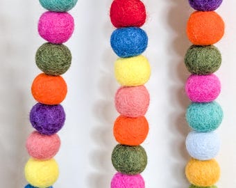 May include: A close-up of three strands of colorful felt ball garlands. The garlands feature a variety of colors, including pink, orange, green, blue, yellow, and purple. The felt balls are arranged in a vertical line, creating a decorative and playful look.