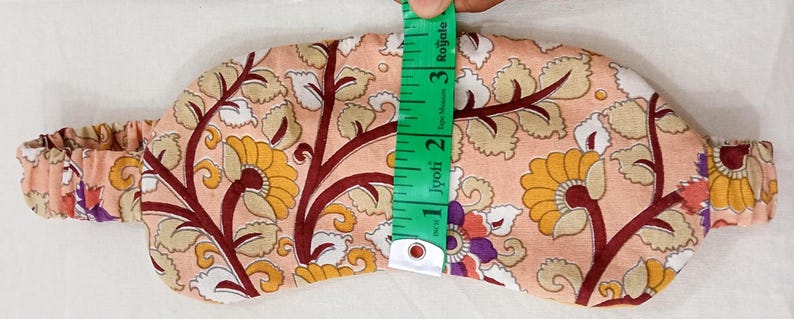 May include: A floral patterned eye mask in peach, yellow, and burgundy. The mask is approximately 3 inches (7.6 cm) wide and has an elastic band. The design features stylized flowers and leaves.