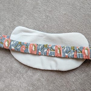 May include: A white, oval-shaped eye mask with a floral patterned headband. The headband features a blue background with pink and green floral designs. The mask is made of a soft, plush material.