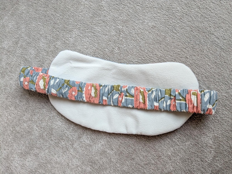 May include: A white, oval-shaped eye mask with a floral patterned headband. The headband features a blue background with pink and green floral designs. The mask is made of a soft, plush material.