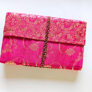 Può includere: Una scatola rettangolare in tessuto rosa acceso con chiusura a patta. La patta e la parte superiore della scatola presentano un motivo broccato floreale dorato. Un cordoncino intrecciato assicura la patta. La scatola è probabilmente utilizzata per riporre oggetti o come oggetto decorativo.