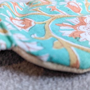 May include: Close-up of a round, floral-patterned fabric item. The fabric features a teal background with white, orange, and gold floral designs. The item has a beige border and a gathered edge.