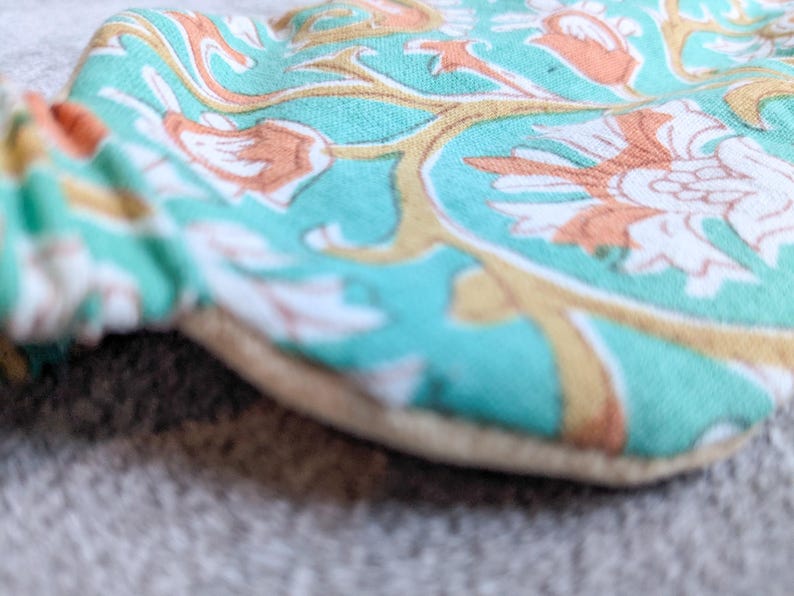 May include: Close-up of a round, floral-patterned fabric item. The fabric features a teal background with white, orange, and gold floral designs. The item has a beige border and a gathered edge.