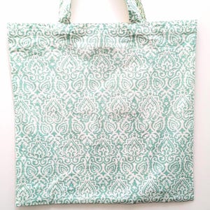 May include: A rectangular tote bag with a repeating white and teal pattern. The bag has two handles made of the same patterned fabric. The pattern is a stylized floral design. The bag is made of a soft fabric.