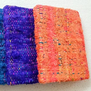 May include: Close-up of three rectangular woven fabric pieces in shades of blue and orange. The fabrics have a textured, handmade appearance with visible threads and a mix of colors, including pink, yellow, and teal.