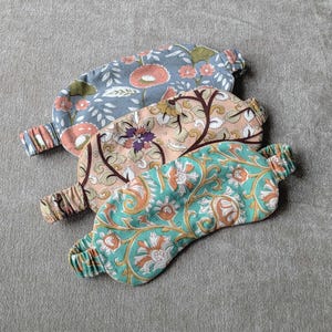 May include: Three floral patterned sleep masks in shades of blue, peach, and teal. Each mask has an elastic band for a comfortable fit. The masks feature various floral designs, including poppies and stylized leaves.