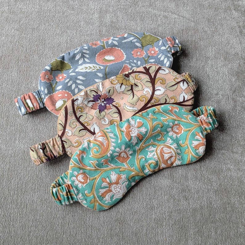 May include: Three floral patterned sleep masks in shades of blue, peach, and teal. Each mask has an elastic band for a comfortable fit. The masks feature various floral designs, including poppies and stylized leaves.