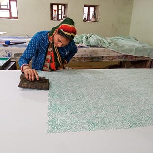 May include: A person using a wooden block to print a green floral pattern onto white fabric. The person is wearing a blue top and a head covering with red, green, and orange stripes. The work is being done on a large table in a workshop.