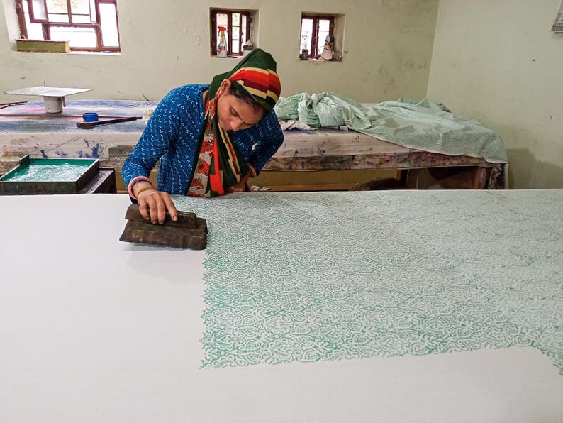 May include: A person using a wooden block to print a green floral pattern onto white fabric. The person is wearing a blue top and a head covering with red, green, and orange stripes. The work is being done on a large table in a workshop.