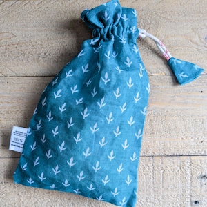 May include: A teal drawstring bag with a white leaf pattern. The bag is made of fabric and has a white drawstring closure with a small fabric tassel. The bag is on a wooden surface.