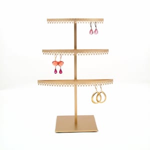 May include: Gold metal earring stand with three tiers. The stand has a square base and a decorative design. There are three pairs of earrings on the stand: one pair of pink teardrop earrings, one pair of pink heart-shaped earrings, and one pair of gold hoop earrings.