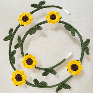 以下が含まれることがあります： ひまわりのデザインが施された、かぎ針編みの充電ケーブル。ケーブルは緑色で、黄色いひまわりと茶色の中心部があります。ケーブルの端は白です。