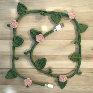 Puede incluir: Una vid verde tejida a ganchillo con hojas y flores rosas, envuelta alrededor de un cable de carga USB blanco. El cable tiene un conector USB-A y Lightning. El diseño está sobre una superficie de madera clara.