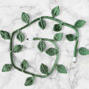 May include: Green crochet USB charging cables designed to look like leafy vines. The cables have a white USB connector at each end. The leaves are a textured, detailed design. The cables are arranged on a marble surface.