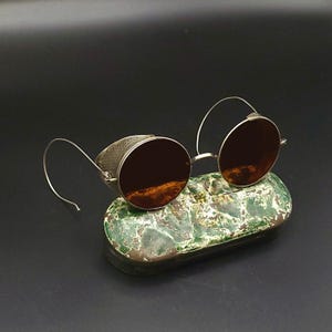May include: Round sunglasses with dark brown lenses and thin silver-toned metal frames. The eyewear features side shields with a perforated pattern and curved wire temples. The glasses are resting on a small, oval-shaped green and white mottled case.