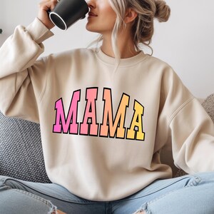 Puede incluir: Sudadera beige con la palabra "MAMA" en letras de bloque degradadas en rosa, naranja y amarillo. La sudadera tiene cuello redondo y mangas largas. La imagen muestra a una persona usando la sudadera.