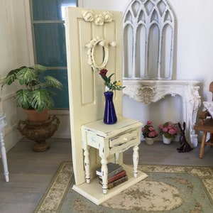 May include: A white painted wooden door with a wreath and floral accents, a small white painted side table with a drawer and turned legs, a blue vase with red and yellow flowers, and a stack of books. The table is on a cream and blue rug.
