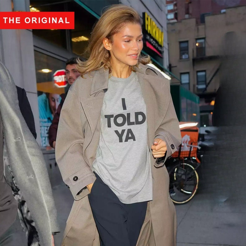 May include: A woman wearing a gray t-shirt with the text "I TOLD YA" printed in black. She is wearing a brown coat and black pants.