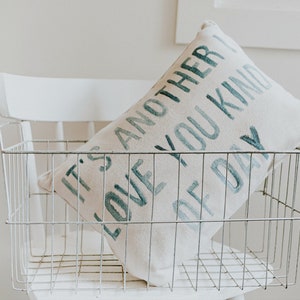 May include: A white pillow with the text "It's another love you kind of day" in teal blue lettering, sitting in a white wire basket.