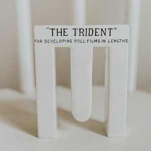 May include: White plastic film developing tank with the text "THE TRIDENT" and "FOR DEVELOPING ROLL FILMS IN LENGTHS."
