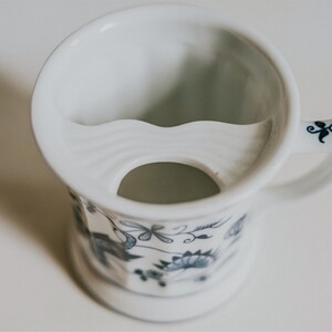Puede incluir: Taza de afeitar de cer&aacute;mica blanca con un patr&oacute;n floral azul y blanco. La taza tiene un reposamustache incorporado y un peque&ntilde;o agujero en la parte inferior para el drenaje.