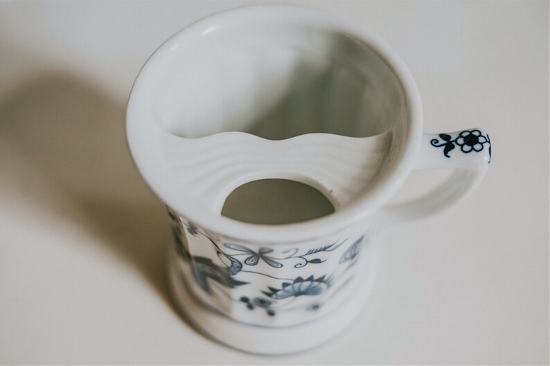 Puede incluir: Taza de afeitar de cer&aacute;mica blanca con un patr&oacute;n floral azul y blanco. La taza tiene un reposamustache incorporado y un peque&ntilde;o agujero en la parte inferior para el drenaje.