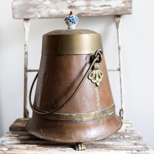 May include: A copper bucket with a brass rim and handle, topped with a lid and a decorative blue and white ceramic knob. The bucket is sitting on a distressed wooden chair.