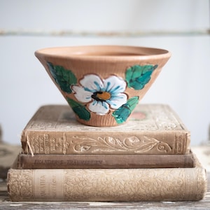 May include: A terracotta bowl with a white flower and green leaves painted on the side. The bowl is sitting on a stack of old books.