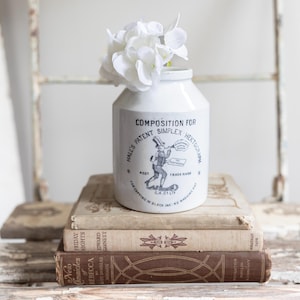 May include: A white ceramic jar with a vintage label and a bouquet of white flowers. The jar is labeled "COMPOSITION FOR SIMPLEX HECTOGRAPH" and is sitting on top of three antique books. The books are stacked on a distressed, white wooden surface.
