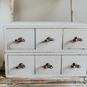 May include: A white wooden drawer organizer with six drawers and antique-style metal pulls. The drawers are stacked in two rows of three.