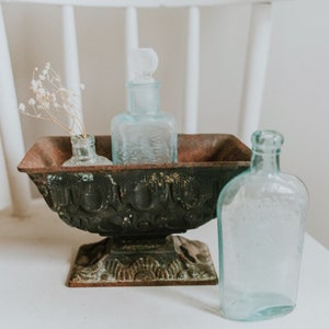 May include: Three clear glass bottles with stoppers, two in a rustic black metal planter with a decorative base, and one on a white surface. The bottles are empty and have a vintage look.