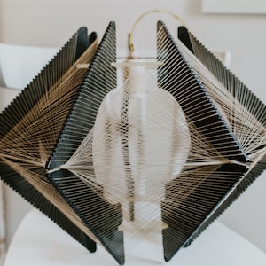 May include: A geometric lampshade made of black wood and string. The lampshade is shaped like a diamond and has a white, round light bulb inside.