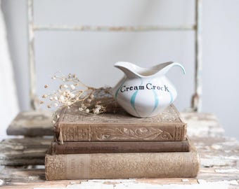May include: A vintage white ceramic cream pitcher with blue and green stripes and the words "Cream Crock" on the side. The pitcher is sitting on a stack of three antique books.