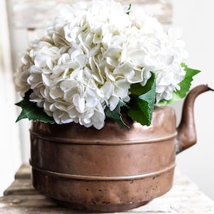 May include: A copper-colored vintage teapot filled with a large arrangement of white hydrangea flowers and green leaves. The teapot sits on a rustic wooden surface, creating a charming, farmhouse-style aesthetic.