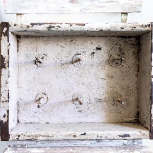 May include: A distressed, rectangular wooden box with a white painted finish and visible wear. The box has metal corner reinforcements and several protruding metal pegs inside. The box is sitting on a white wooden chair.
