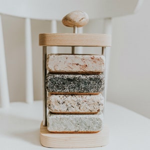 May include: A set of four coasters made of different types of stone, stacked on a wooden stand with a stone top. The coasters are in a variety of colors, including gray, white, and brown.