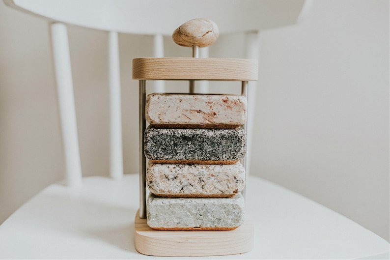 May include: A set of four coasters made of different types of stone, stacked on a wooden stand with a stone top. The coasters are in a variety of colors, including gray, white, and brown.