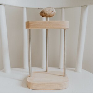 May include: A wooden stand with three metal rods and a round wooden top. A small, light brown rock sits on the top of the stand.