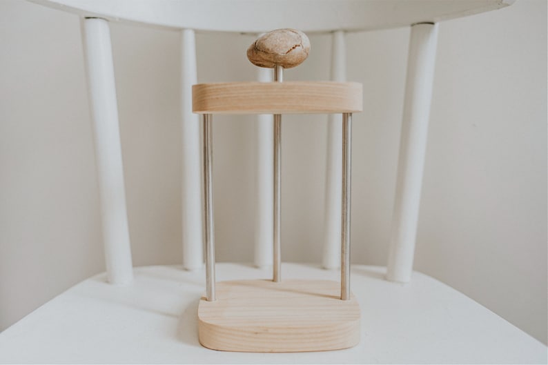 May include: A wooden stand with three metal rods and a round wooden top. A small, light brown rock sits on the top of the stand.
