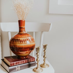 May include: A brown and orange ceramic vase with a geometric design, filled with dried white flowers, sits on a stack of three books with gold candle holders on a white chair.